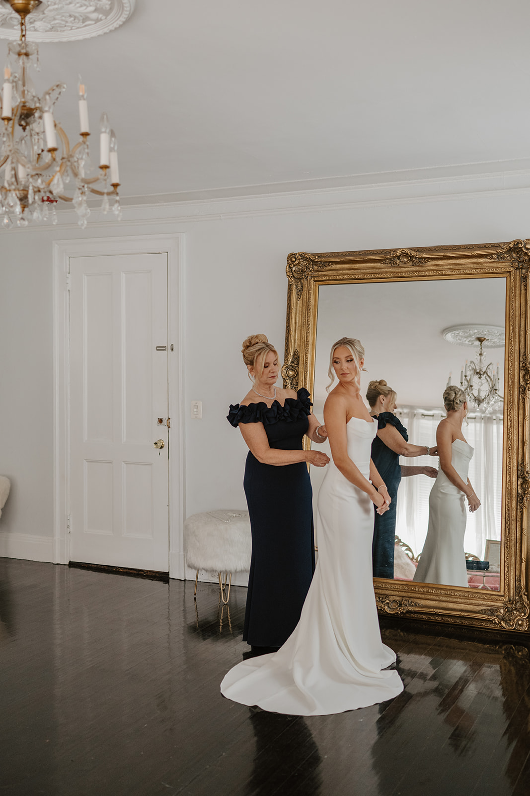 Mother of the bride zips up the bride's elegant white gown in front of a grand gilded mirror.